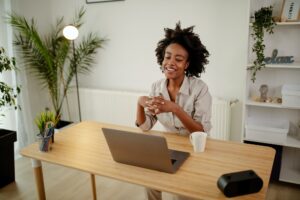 Black smiling businesswoman using laptop, drinking a coffee and having conference call from home.